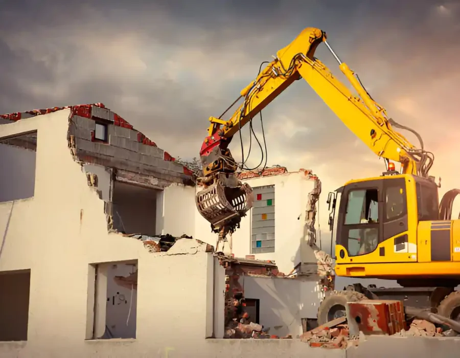 Yellow excavator using grapple attachment to dismantle white building