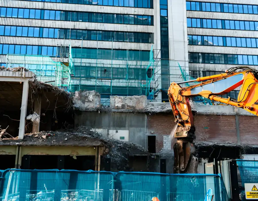 Yellow excavator dismantling concrete structure near modern glass office building