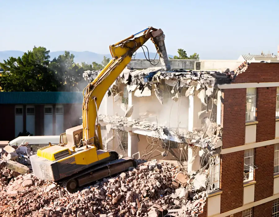 Yellow excavator demolishing multi-story brick building, debris cleanup in progress