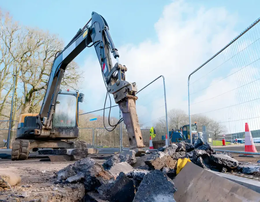 Excavator with hydraulic breaker attachment breaking concrete at fenced demolition site with safety cones