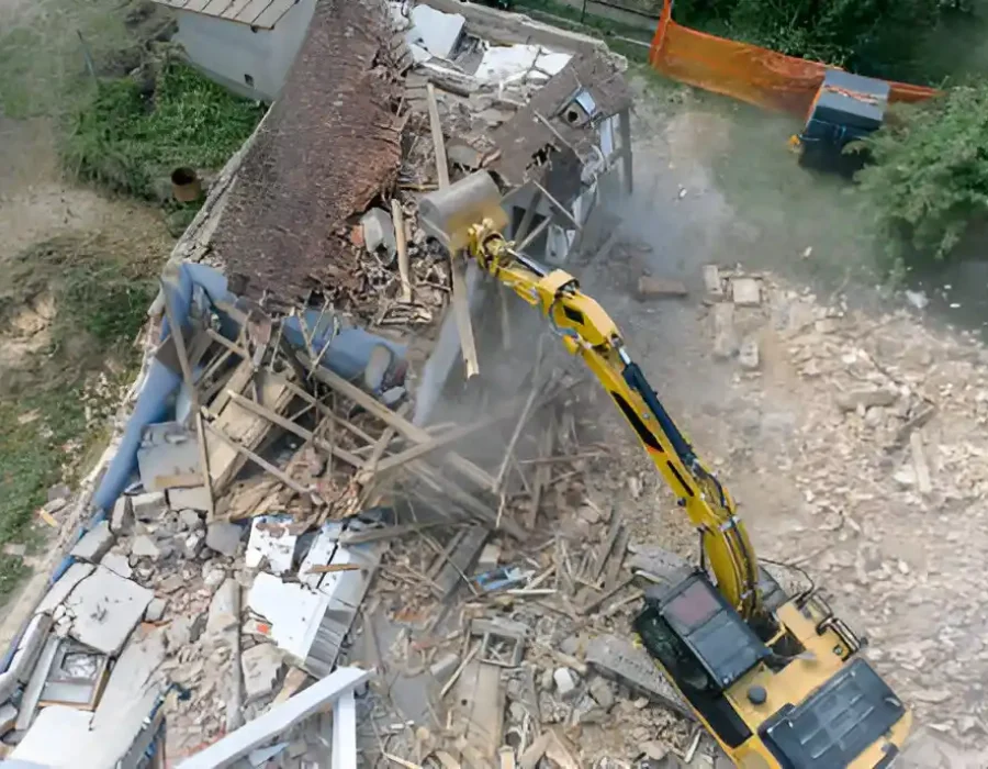 Yellow excavator dismantling wooden building, debris scattered across demolition site