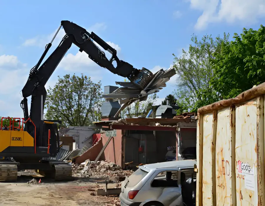 Black excavator destroying small building, debris and storage container in demolition site