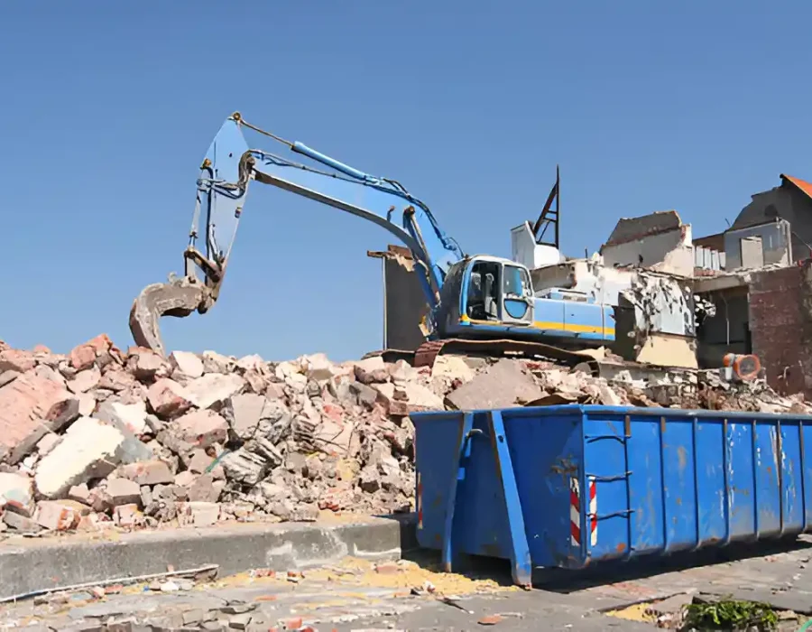 Blue excavator loading rubble into dumpster, residential building demolition site