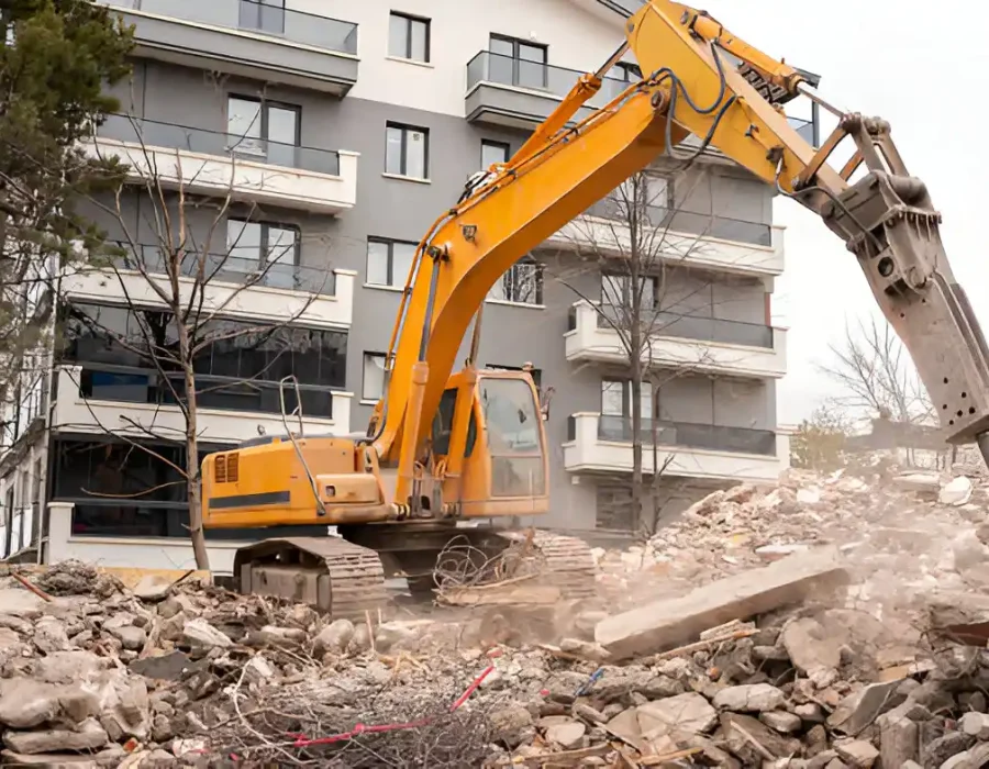 Yellow excavator dismantling concrete building, debris scattered near modern apartments