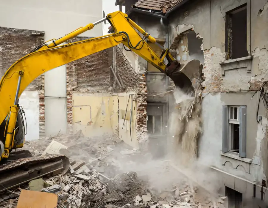 Yellow excavator breaking down old brick building with dust and debris