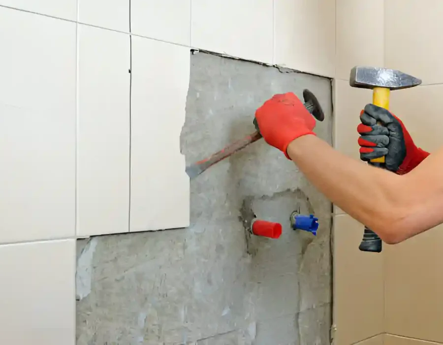 Worker removing bathroom tiles with hammer during demolition project, wearing safety gloves for tile removal