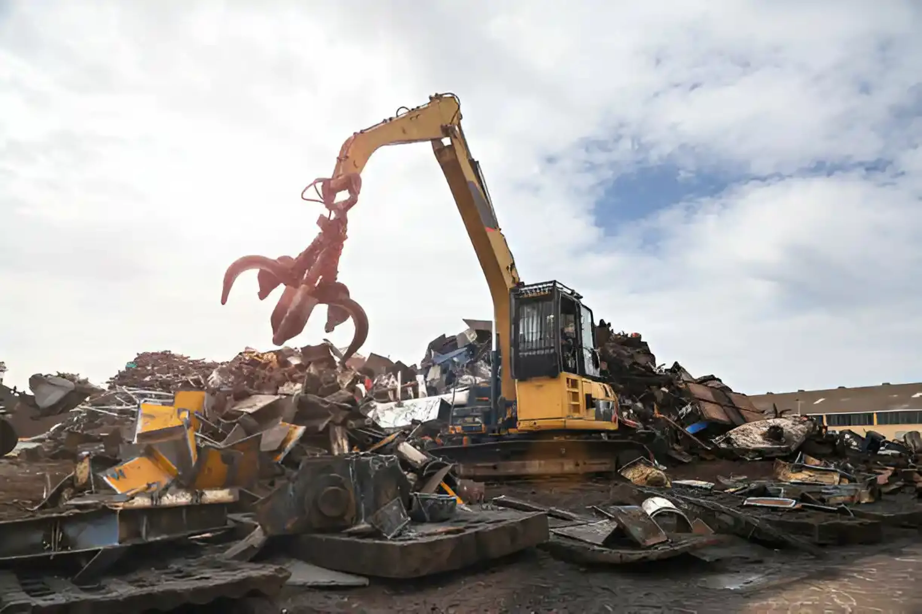 Yellow excavator with hydraulic grapple sorting metal waste at industrial recycling yard under cloudy sky