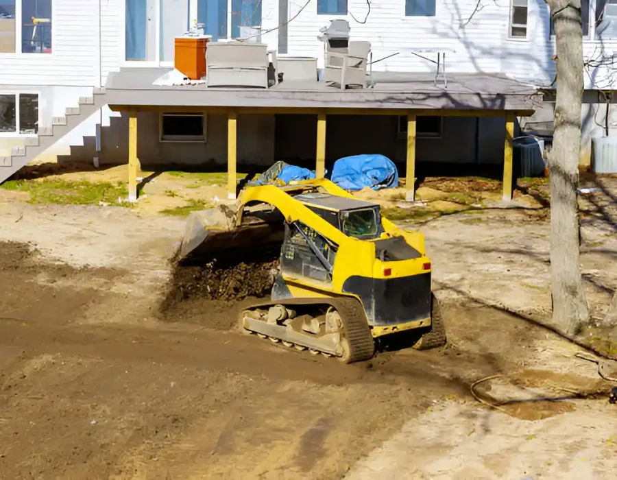 Excavator leveling soil for debris removal and site preparation near house foundation