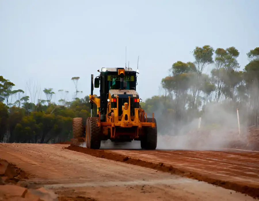 Industrial motor grader preparing construction site with dust cloud and eucalyptus trees in background