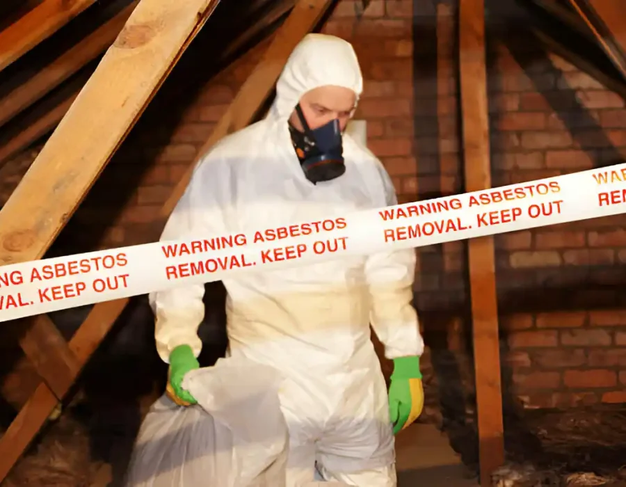 Worker in hazmat suit conducts asbestos removal, marked by warning tape in the attic.
