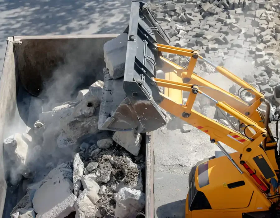 Yellow loader dumping concrete demolition debris into industrial waste container with dust control