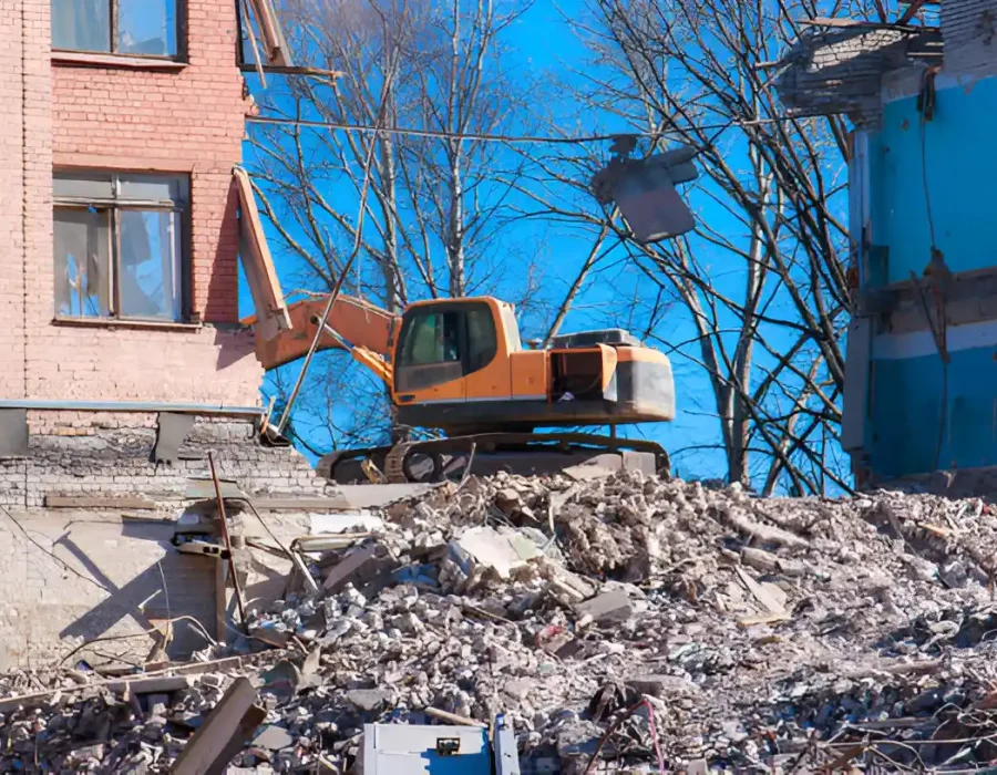 Orange excavator on debris pile demolishing brick structure against bright blue sky