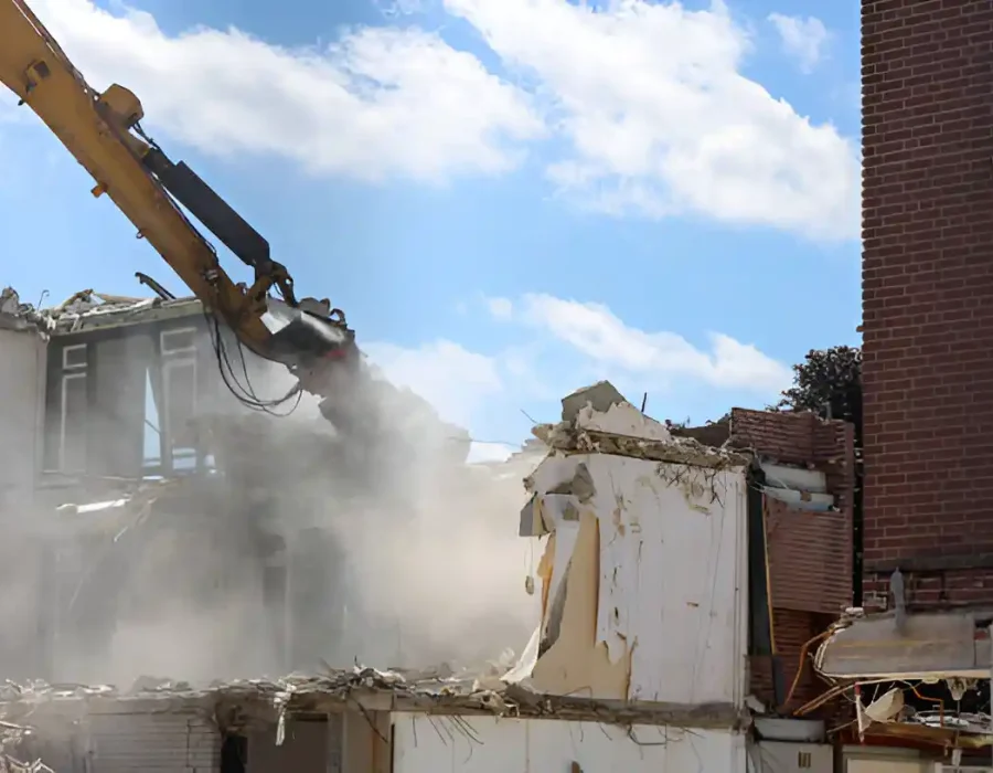 Excavator demolishing white building with active dust suppression in urban environment