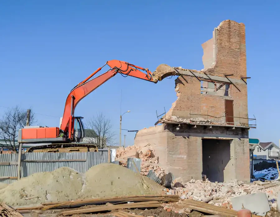 Orange excavator demolishing tall brick chimney structure during industrial site preparation