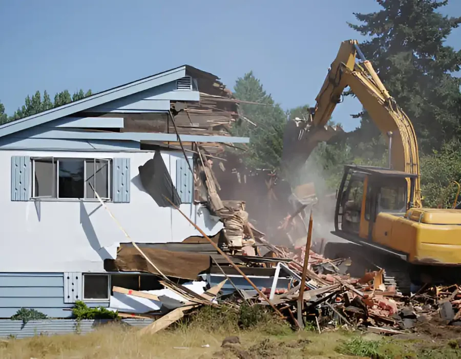 Yellow excavator dismantling white house, debris scattered, professional demolition service