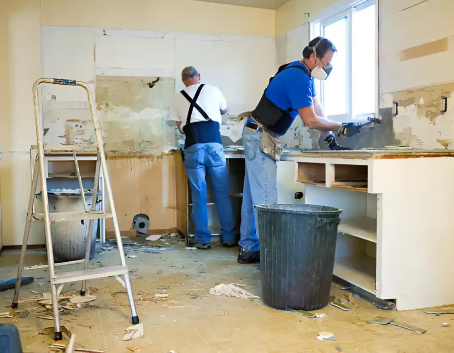 Two workers removing kitchen cabinets, debris on floor, ladder and trash can visible