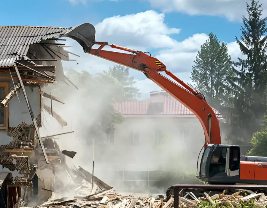 Orange excavator demolishing white house, dust clouds, roof collapse in progress