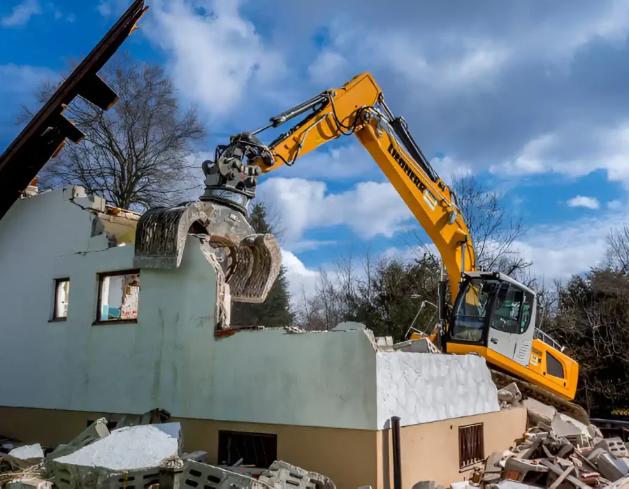 Yellow excavator with grapple crushing white house, debris removal in progress