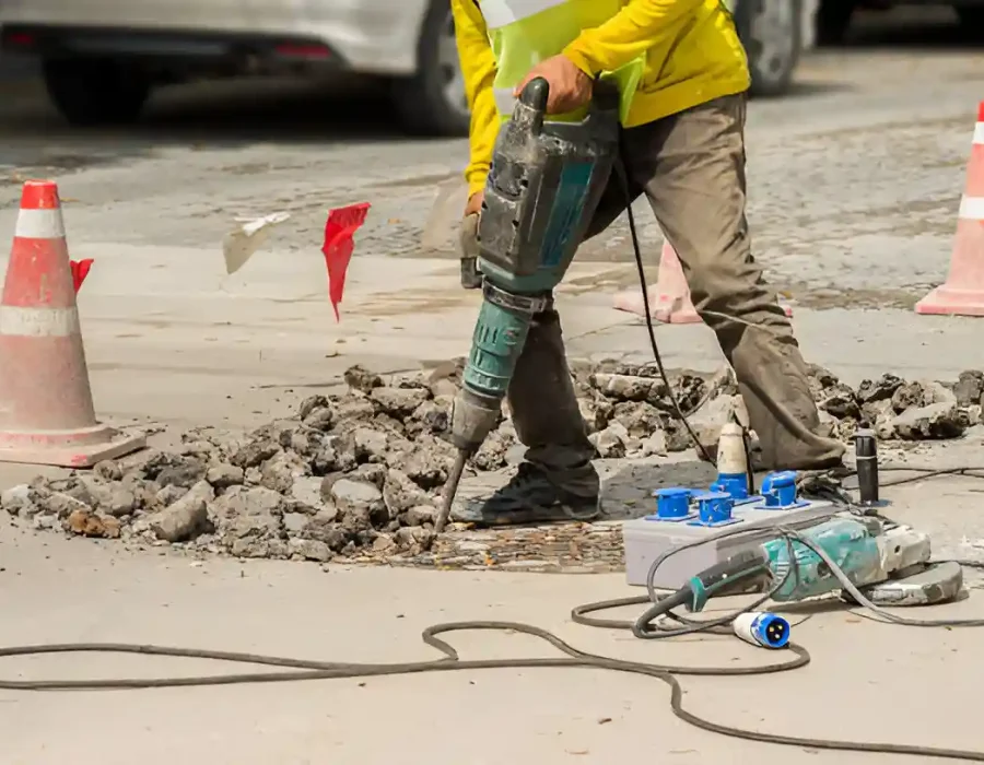 Worker using jackhammer for concrete demolition with safety cones, debris removal and site preparation equipment