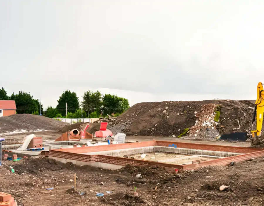 Construction site showing a building foundation, excavator, and piles of dirt and debris.
