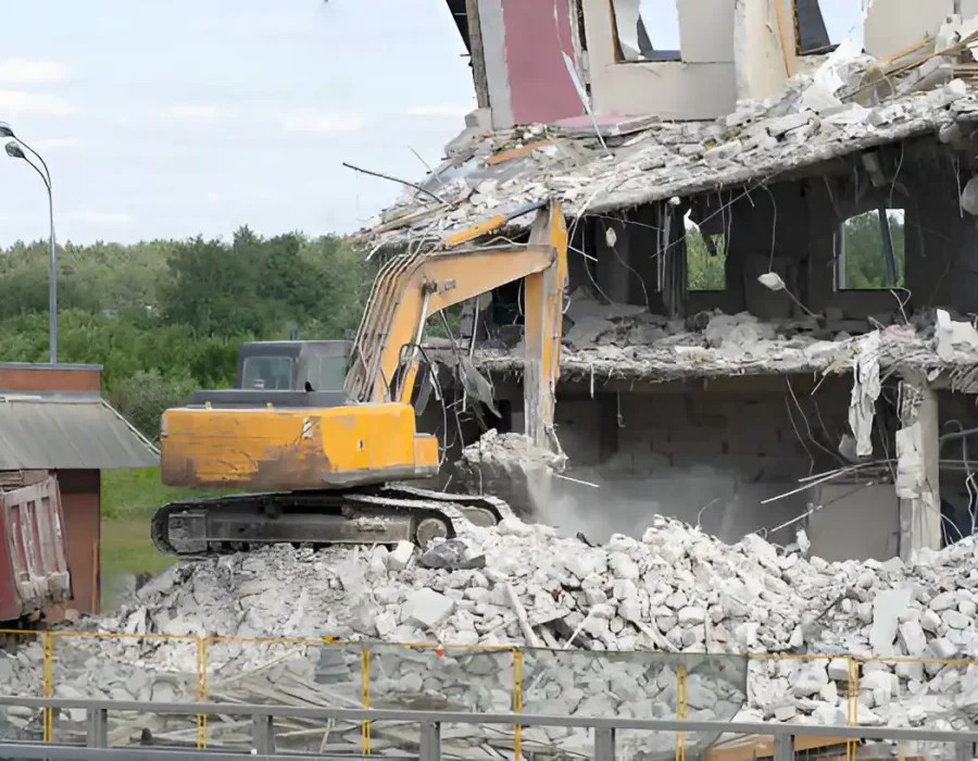 Yellow excavator tearing down a concrete building with rubble and debris scattered around.