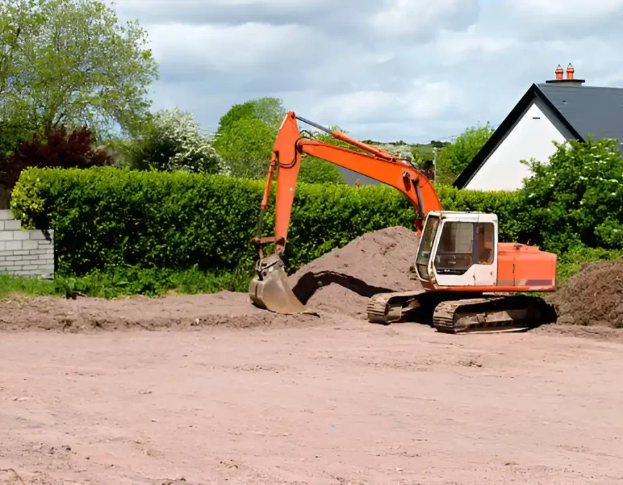 Orange excavator moving dirt near residential house with green hedge background