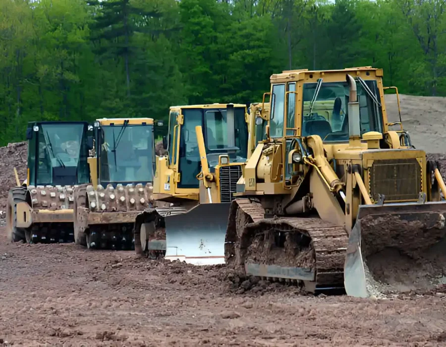 Multiple yellow bulldozers and compactors lined up on dirt terrain with forest background