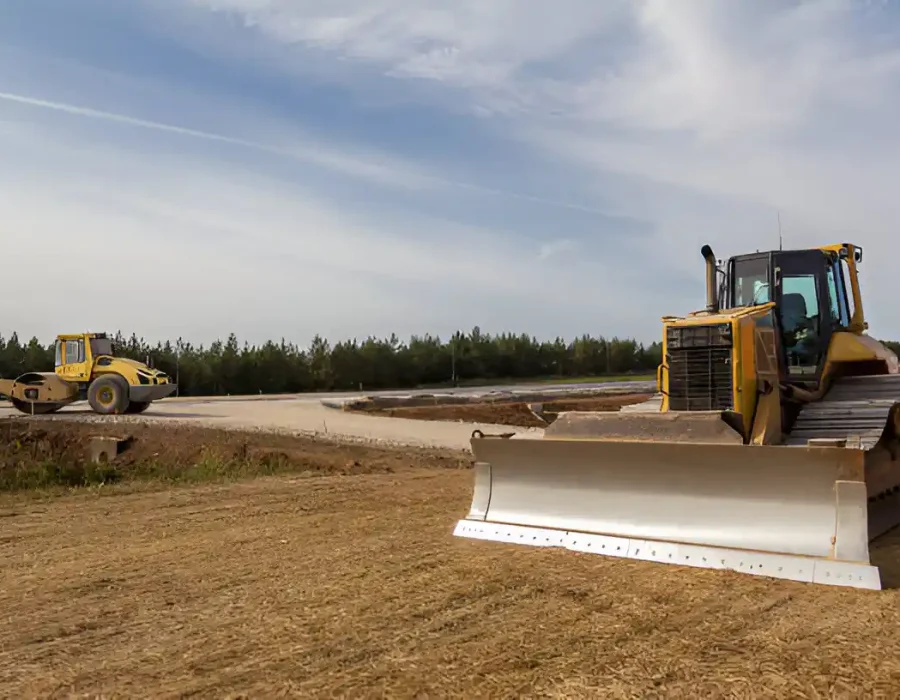 Bulldozer and roller compacting and leveling soil on a large open site near a forested area.