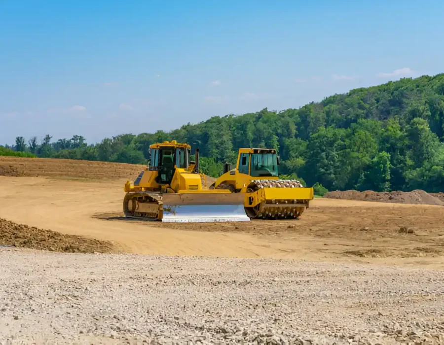 Two yellow compactors preparing construction site with forested green hills in background