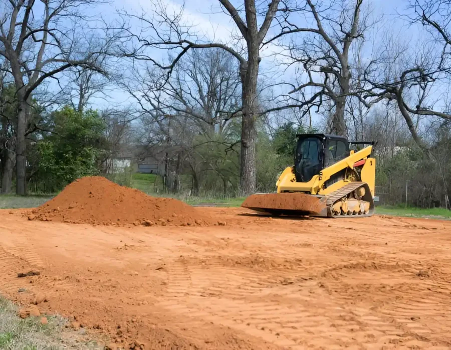 Yellow skid steer machine moving red soil pile near bare trees in open landscape