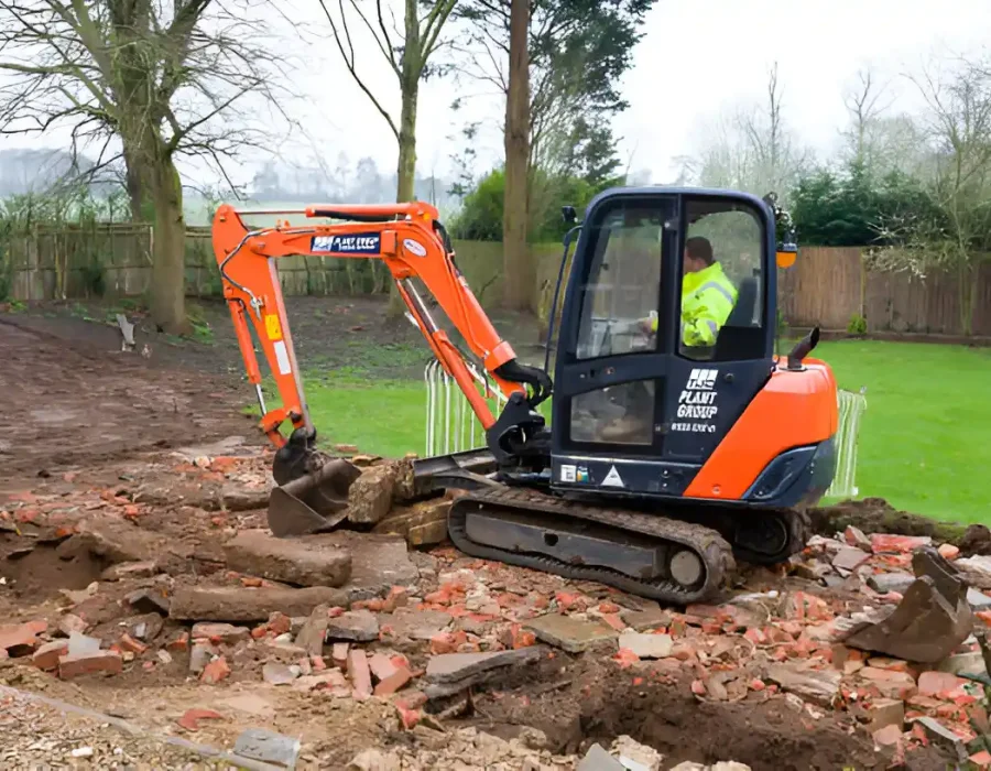 Orange mini excavator removing brick rubble in backyard with green lawn and trees