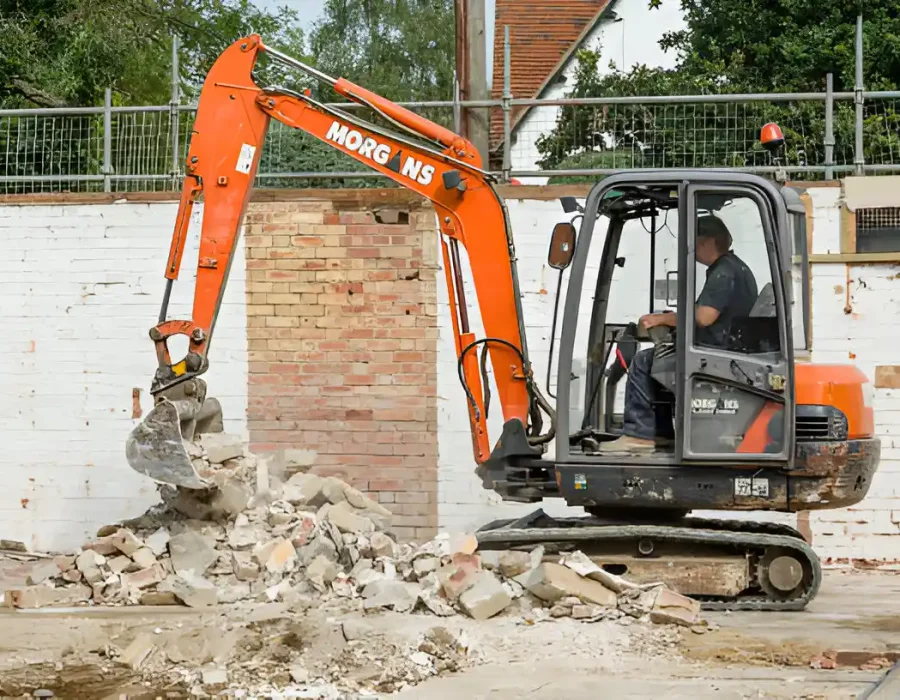 Orange excavator breaking down brick wall with concrete rubble at construction site