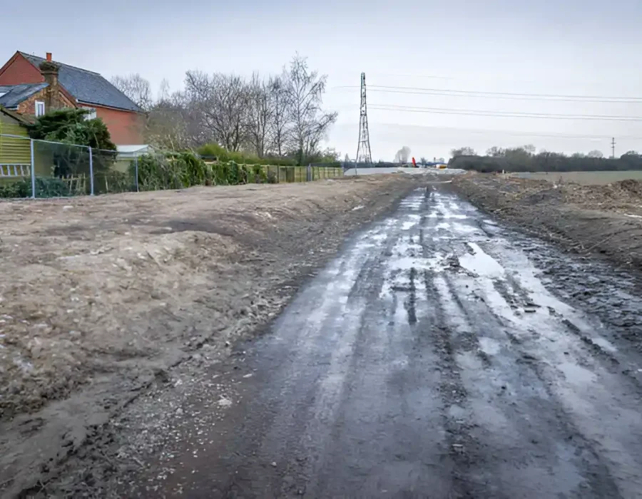 Muddy construction site with wet ground, residential buildings, and power lines in background