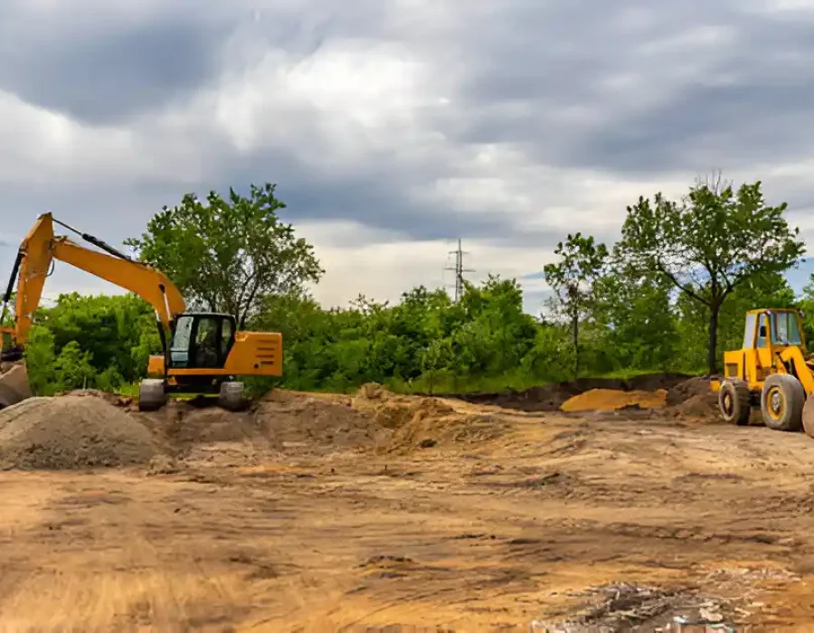 Excavator and loader working on site preparation with dirt piles and trees in the background.