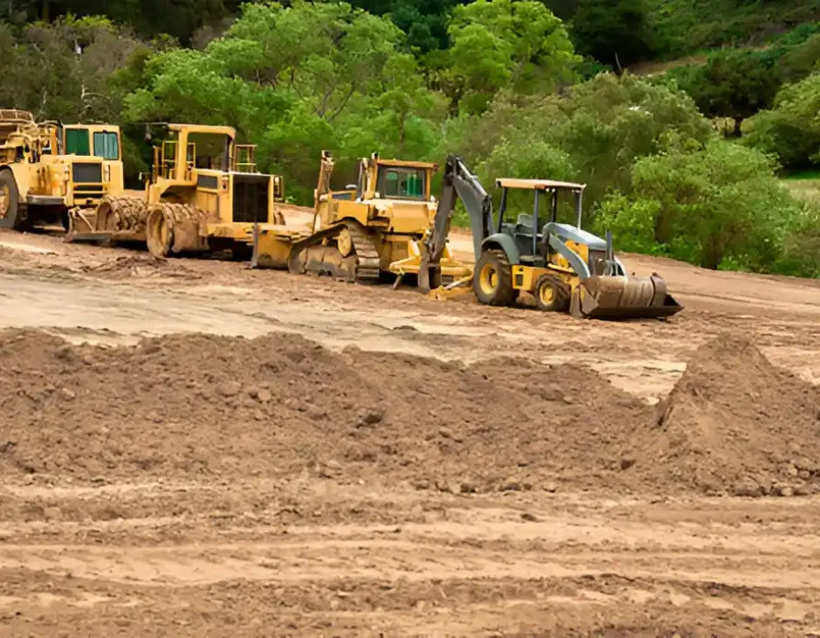 Multiple yellow construction machines lined up on dirt site with green trees