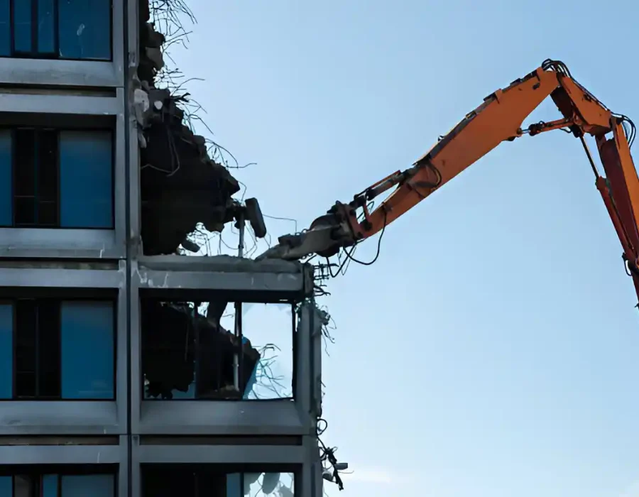 Orange demolition crane removing glass facade panels from modern office building during controlled demolition