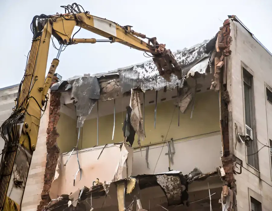 Yellow excavator performing controlled demolition of building exterior wall and structural elements