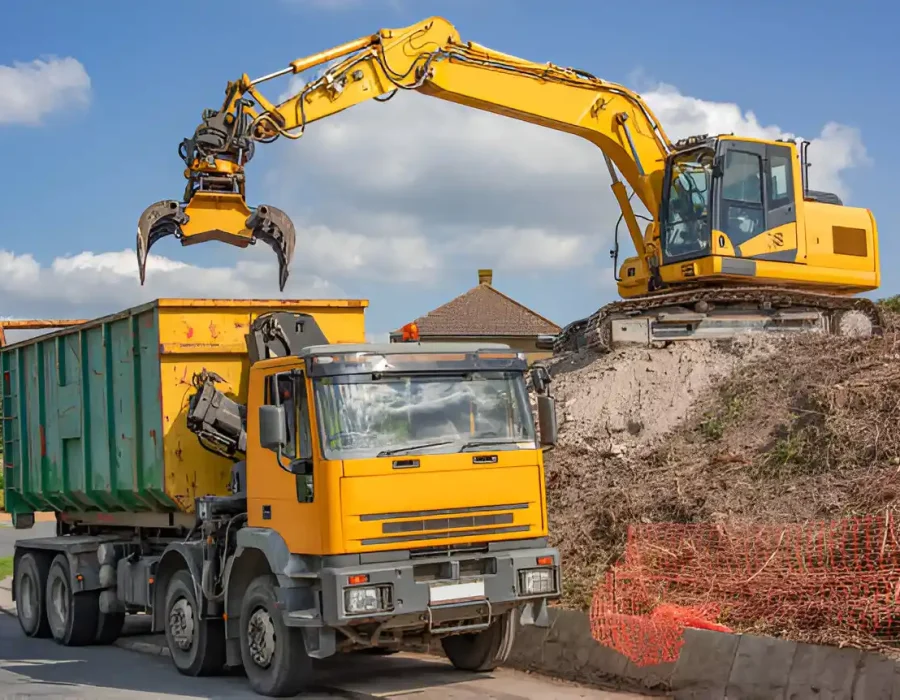 Yellow excavator with grapple loading construction debris into roll-off container truck at worksite