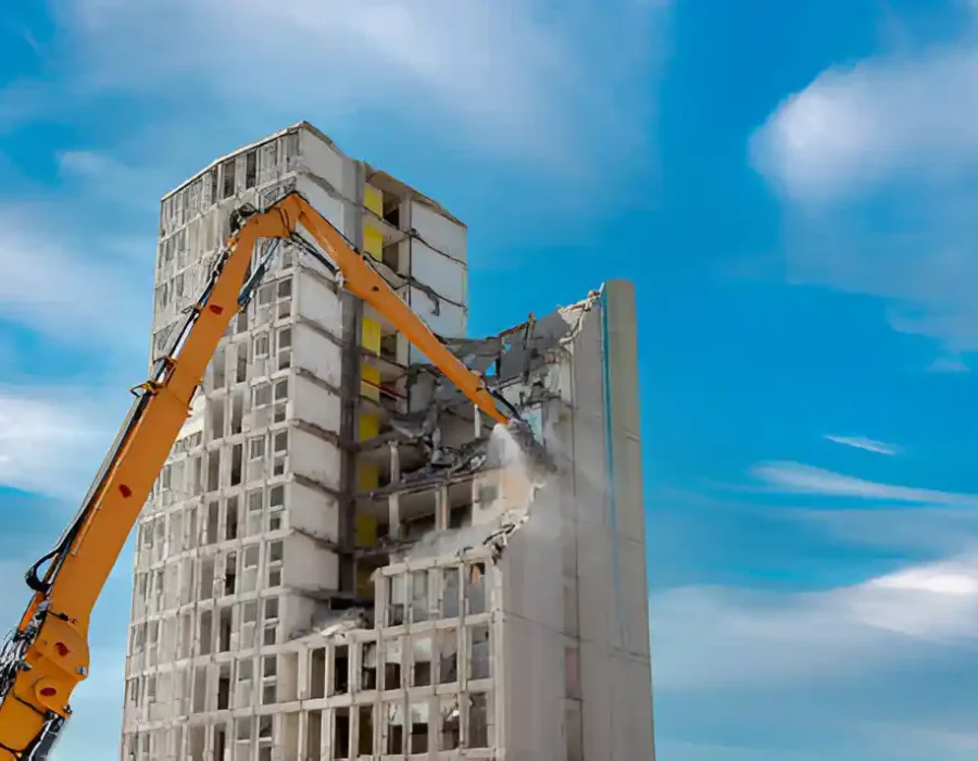 Orange excavator dismantling white multi-story building against blue sky backdrop