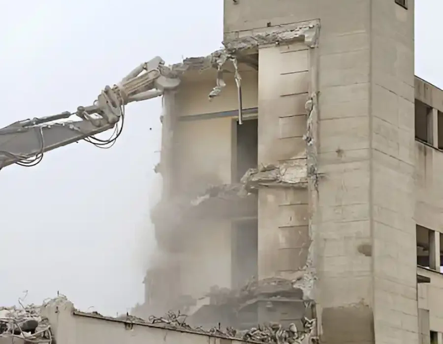 Gray excavator arm demolishing multi-story concrete building with dust clouds