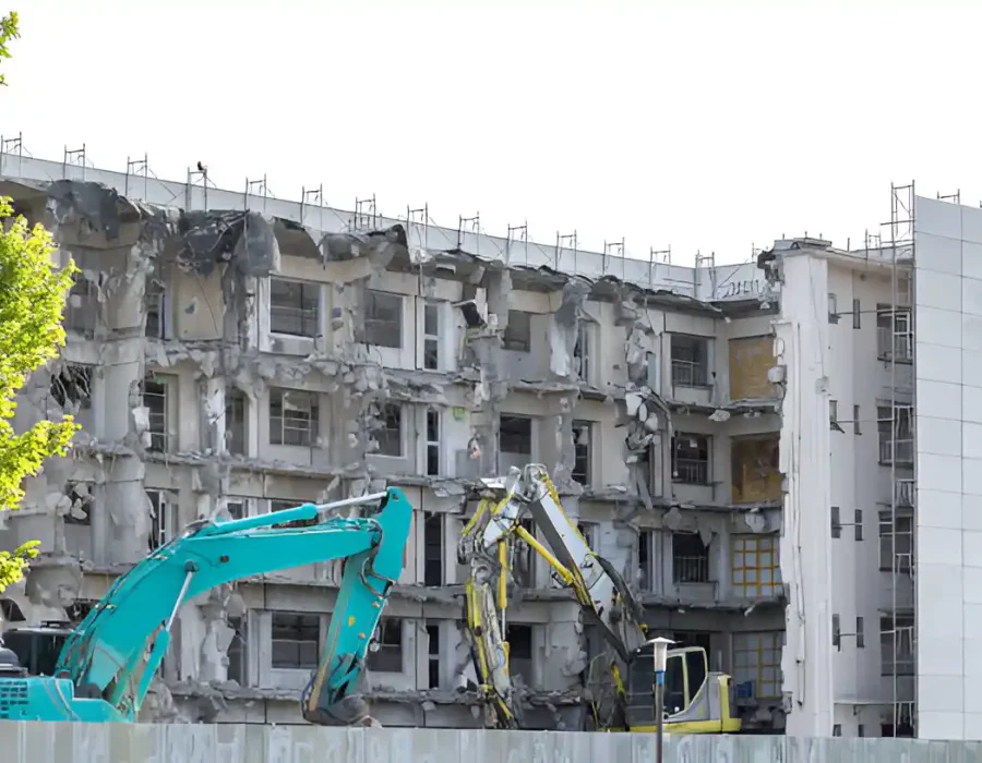 Teal and yellow excavators dismantling white concrete apartment complex