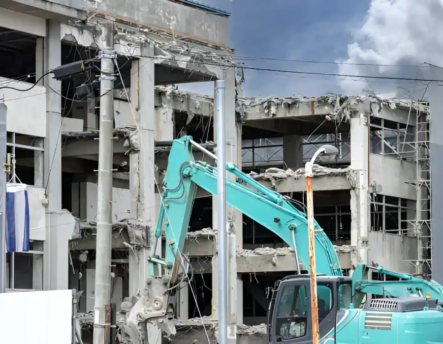 Teal excavator dismantling white multi-story commercial building with scaffolding