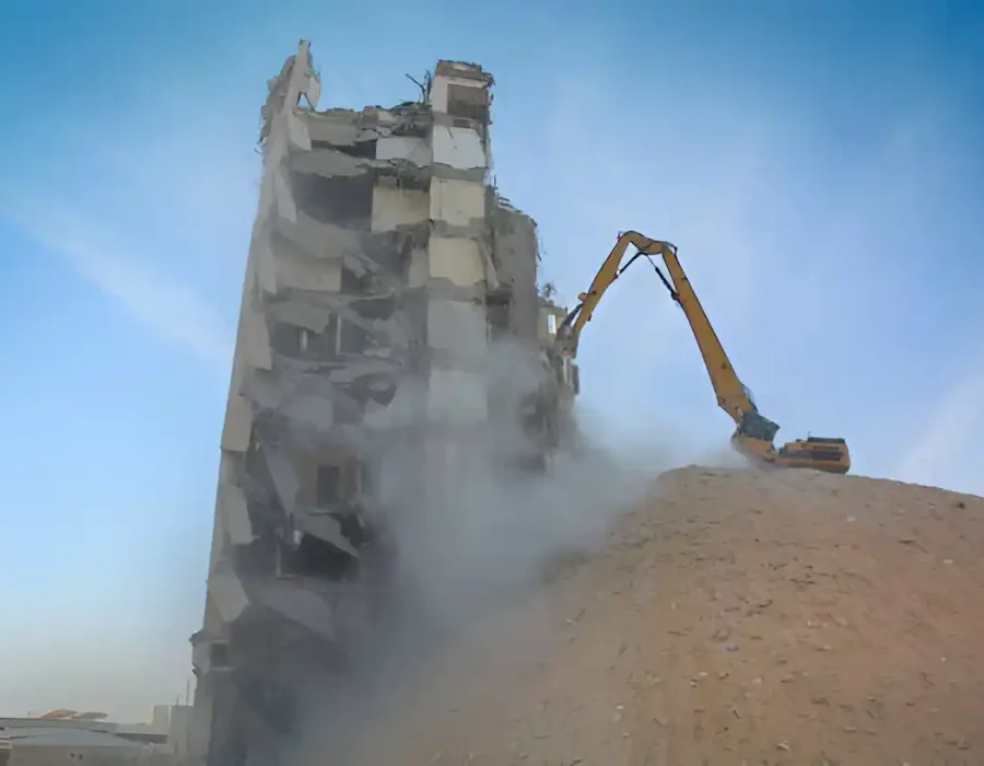 Long-arm excavator dismantling white concrete building with dust cloud