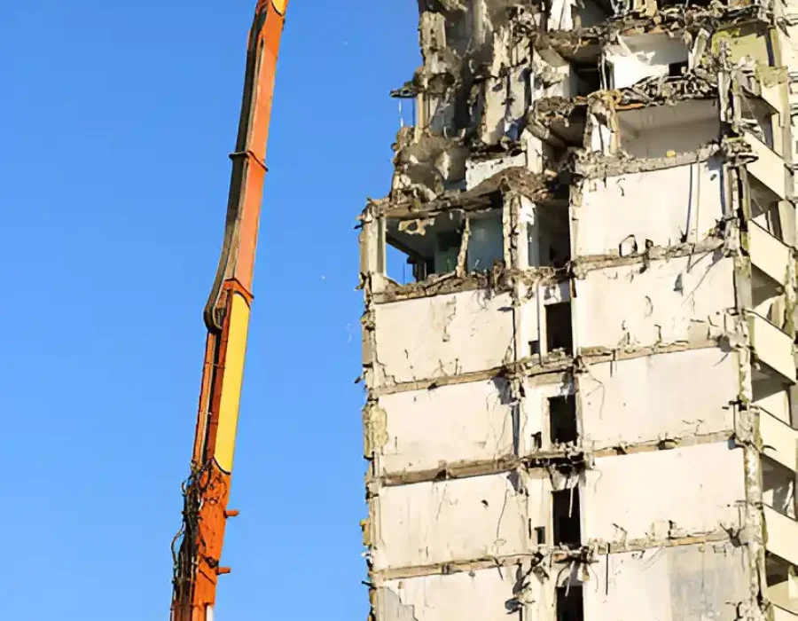 Orange long-reach demolition machine positioned on debris mound targeting tall building structure