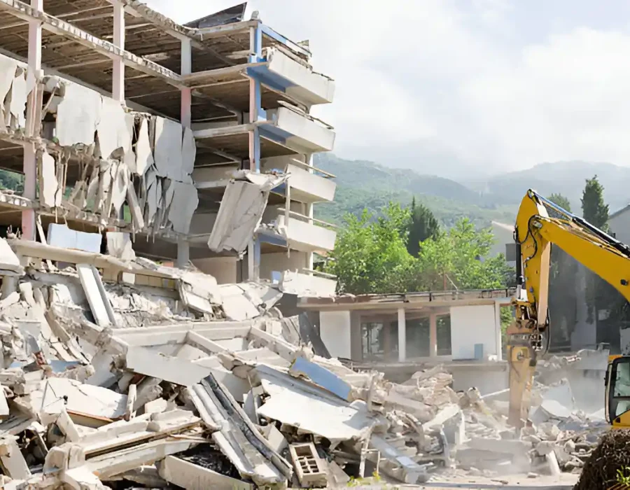 Yellow excavator dismantling concrete apartment complex, massive debris pile in foreground