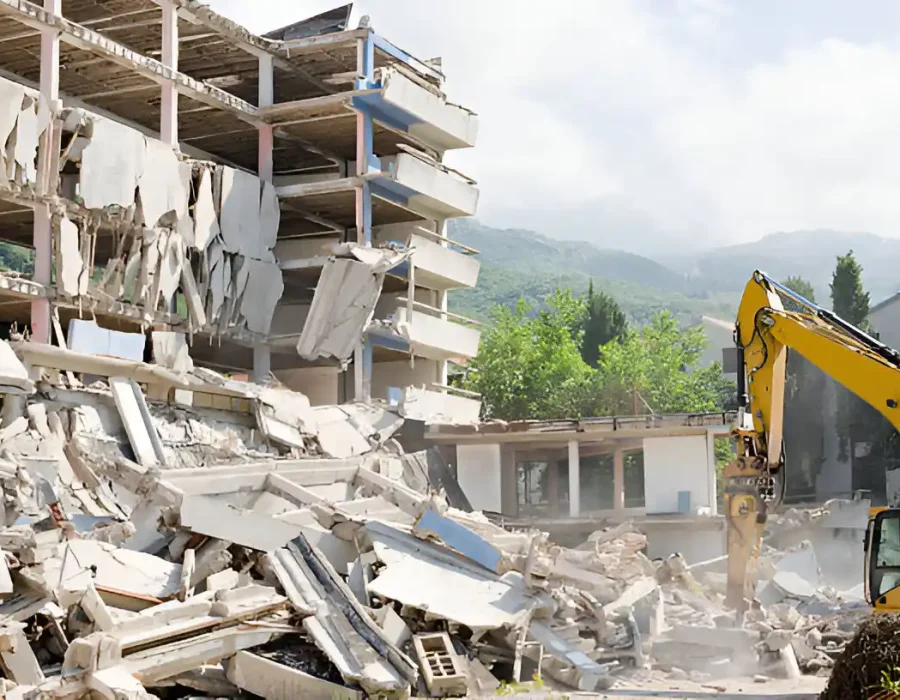Excavator demolishing concrete structure with mountain backdrop, debris removal in progress