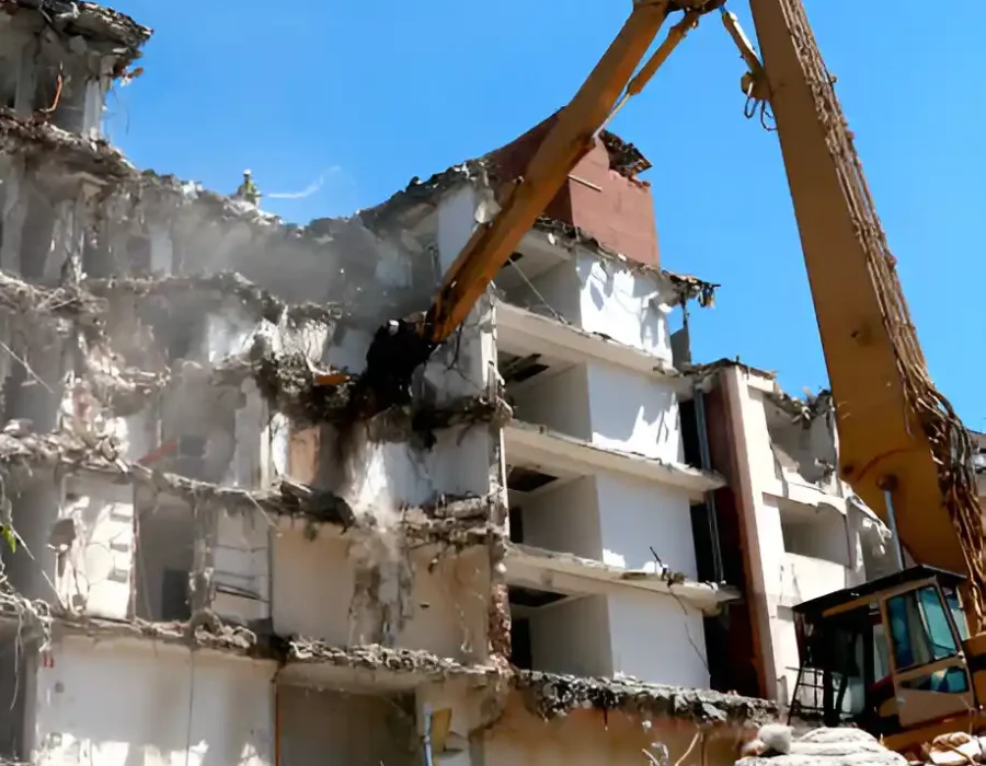 Long-arm excavator dismantling white concrete building, massive debris pile below