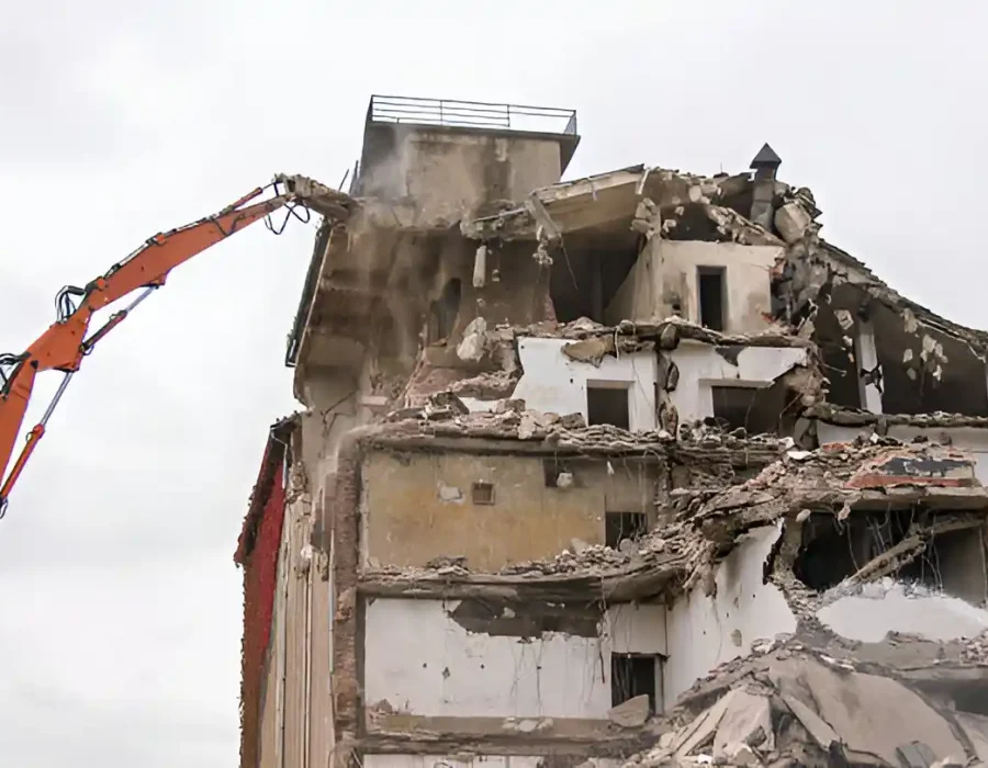 Orange excavator dismantling multi-story white building, structural damage visible