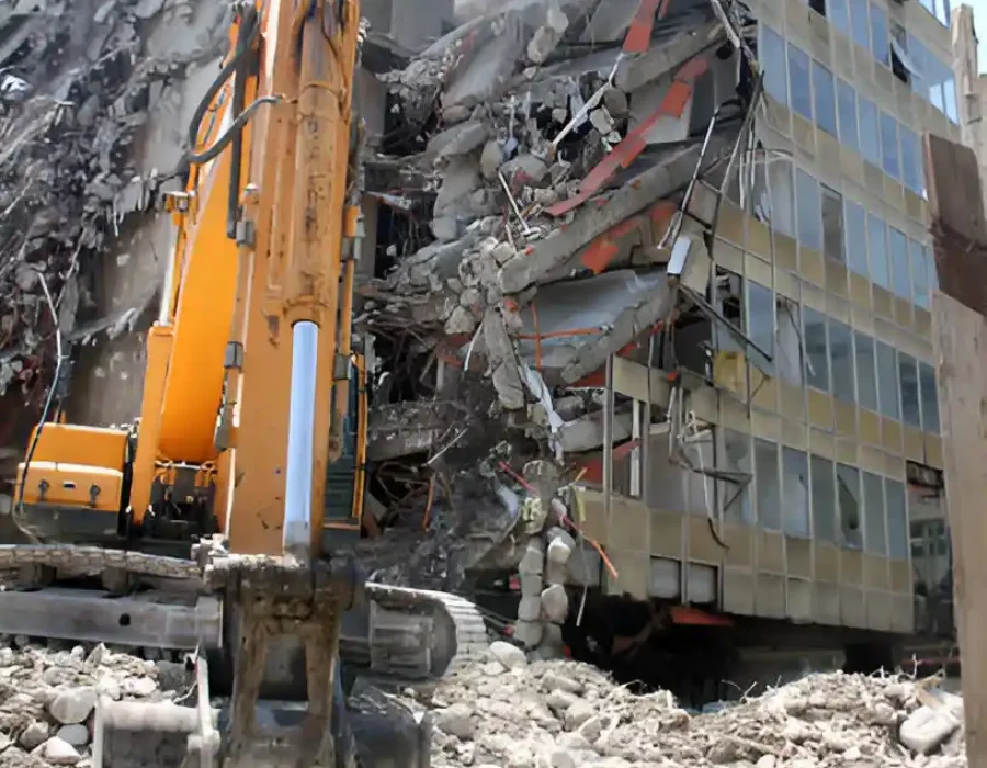 Orange excavator dismantling multi-story glass and concrete building, massive debris