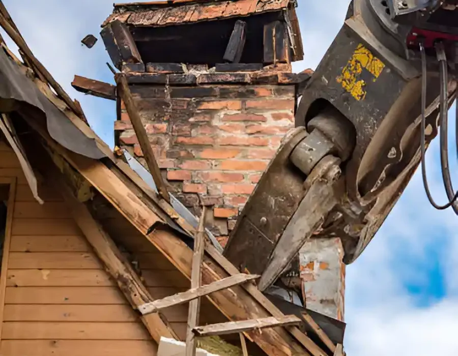 Excavator claw tearing down a brick chimney and roof during a residential demolition project.